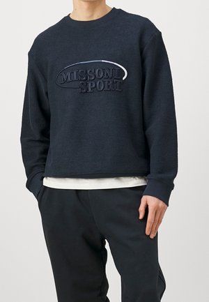 Mann trägt schwarzen, strukturierten Langarm-Sweatshirt mit "MISSONI SPORT" auf der Brust bestickt und schwarze Hose, Hände entspannt an den Seiten.