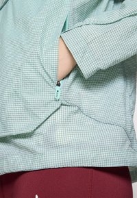Light blue checkered jacket with a zipper pocket and loose fit. Smooth texture and elbow-length sleeves; worn with burgundy leggings.
