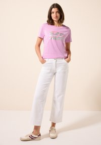 T-shirt rose à manches courtes avec un design graphique argenté, associé à un pantalon blanc évasé et des baskets beiges avec des accents bordeaux.