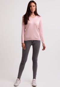Světle růžový dlouhý svetr s výstřihem do V, vyrobený z měkkého materiálu, v kombinaci se šedými skinny džíny a bílými teniskami. Jednoduchý a přiléhavý design.