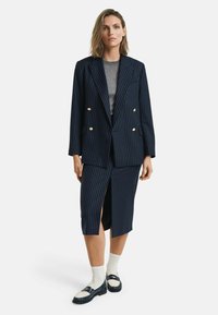 Blazer blu navy a righe con bottoni dorati, abbinato a una gonna midi coordinata con spacco laterale, top grigio e mocassini con calzini bianchi.