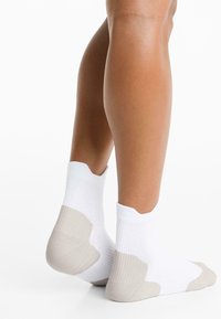 Lábak fehér bokazokniban, bézs sarok- és lábujjrésszel, egy fehér háttér előtt állva.