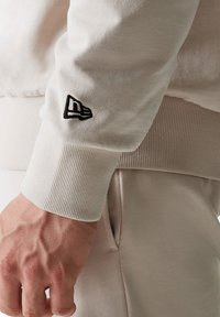 Felpa beige chiaro con polsini a coste. Presenta un piccolo logo nero ricamato sulla manica. Materiale morbido con una texture liscia.