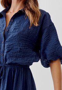 Mörkblå knapplistad klänning med en texturerad, rynkad tyg. Har en krage, korta ärmar och en justerbar dragsko i midjan.