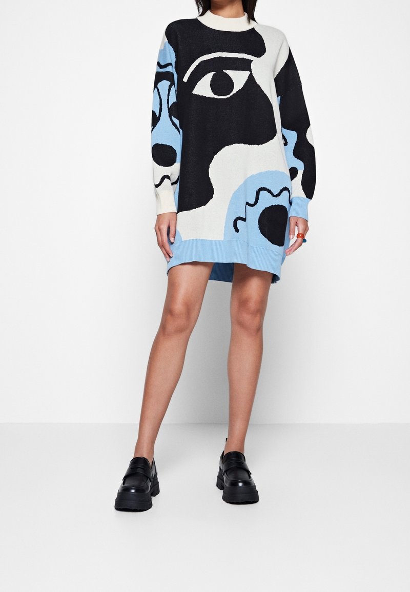 Femme portant une robe-pull oversize noire, blanche et bleue avec des motifs abstraits de visages et des chaussures à plateforme noires épaisses.