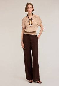 Top beige in seta con scollatura arricciata, abbinato a pantaloni a gamba larga color marrone. I gioielli presentano pannocchie nere e dorate. Tessuto dalla consistenza liscia.
