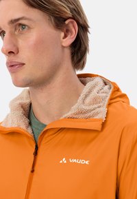 Oranje jas met een met fleece gevoerde capuchon, gladde buitenkant en zwarte rits. Heeft een wit "VAUDE" logo op de borst.