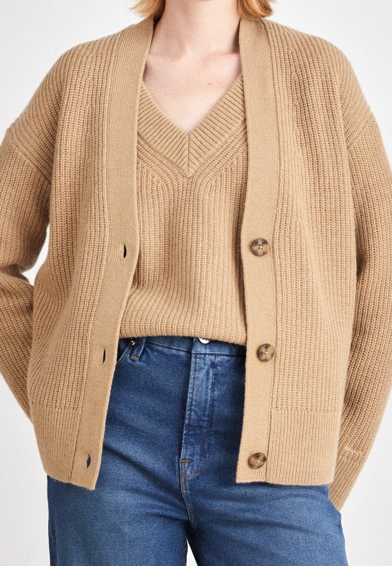 Femme portant un pull en maille beige à col en V superposé d'un cardigan assorti à boutons et un jean bleu taille haute, les mains derrière le dos.