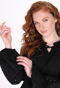 Blusa nera in tessuto testurizzato con motivi a pois, maniche ampie a sbuffo con polsini elastici e scollatura regolabile con cordoncino.