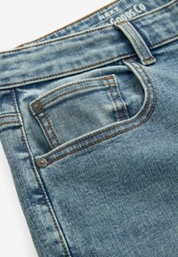 Jeans de mezclilla azul desvanecido con un bolsillo frontal, costuras visibles en naranja y remaches metálicos. Tela texturizada con un ligero efecto de lavado.
