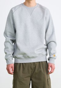 Mann in einem hellgrauen langärmeligen Sweatshirt und olivgrünen Cargo-Hosen, der mit entspannten Händen an den Seiten steht.