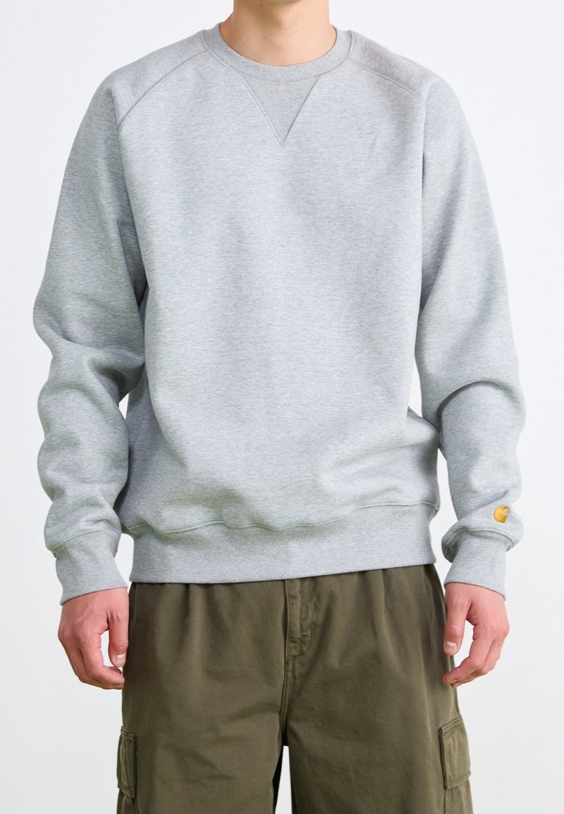 Mann in einem hellgrauen langärmeligen Sweatshirt und olivgrünen Cargo-Hosen, der mit entspannten Händen an den Seiten steht.