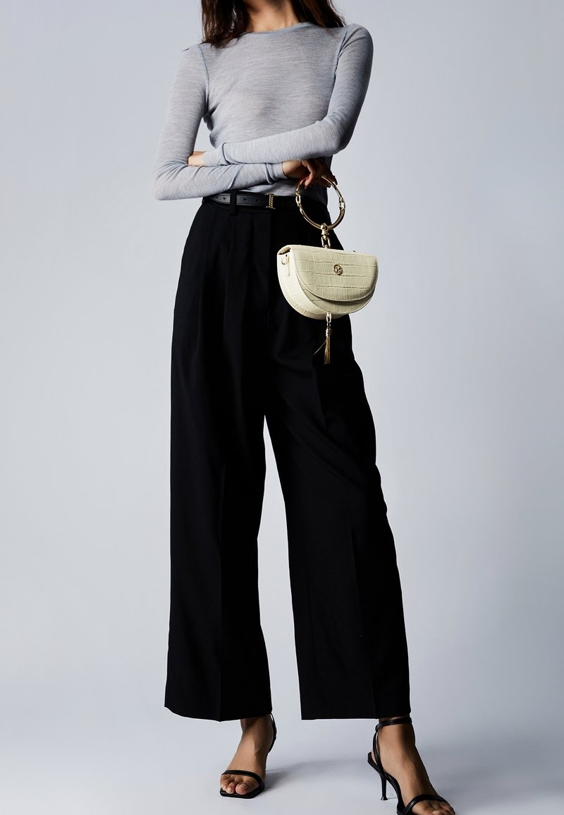 Femme debout, les bras croisés, portant un haut gris à manches longues, un pantalon large noir, des talons noirs et tenant un petit sac à main beige avec une anse en anneau doré.