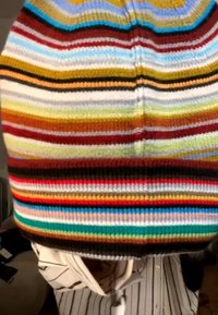 Person trägt eine mehrfarbige, horizontal gestreifte Strickmütze und ein weißes Hemd mit dünnen schwarzen vertikalen Streifen drinnen.