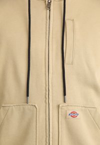 Sweatshirt bege com fecho e cordões pretos, três bolsos e um pequeno logótipo colorido da Dickies no bolso inferior direito.