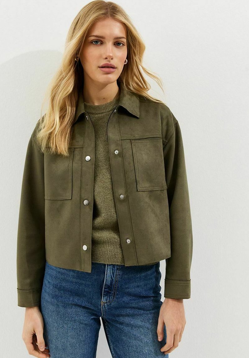 Mujer rubia con chaqueta abotonada verde oliva, suéter de punto y jeans azules, de pie frente a una pared blanca lisa.