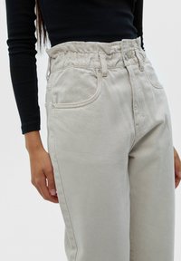 Femme porte un jean beige taille haute avec une ceinture élastique et des poches devant, associé à un haut noir à manches longues.