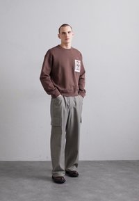 Sweatshirt castanho de gola redonda com um patch gráfico, combinado com calças cargo cinza-claro com bolsos laterais e um corte descontraído. Sapatos escuros.