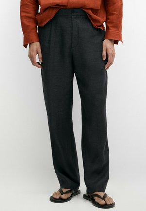 Uomo che indossa pantaloni larghi grigio scuro, infradito in pelle nera e una camicia a maniche lunghe color ruggine con le mani rilassate lungo i fianchi.