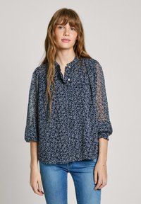 Marinebluse mit floralem Muster und transparenten Ärmeln, Rüschensaum und Knopfdetails. Aus leichtem Stoff und lockerer Passform, kombiniert mit blauen Jeans.