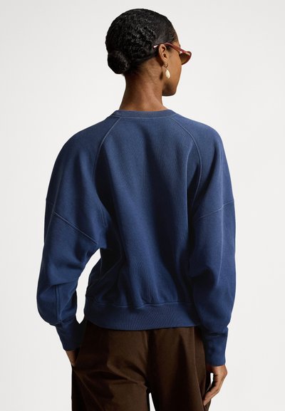 Marineblå genser med avslappet passform, ribbet halslinning og raglanermer. Myk tekstur, uten synlige mønstre, kombinert med brune bukser.