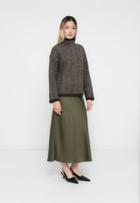 Žena u smešnom i bež prugastom turtleneck puloveru, maslinastozelenoj midi suknji i crnim špicastim slingback cipelama stoji uz bijelu pozadinu.