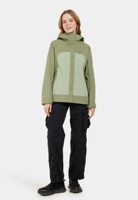Veste légère verte avec capuche, présentant un design texturé et des panneaux contrastants vert clair, associée à un pantalon cargo noir et des chaussures.