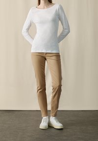 Haut à manches longues blanc avec un col rond, associé à un pantalon beige slim-fit et des baskets blanches avec des semelles crème. Tissu simple et texturé.