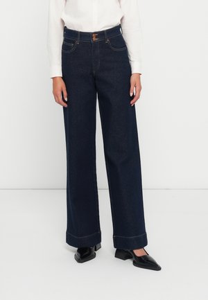 Jeans en denim bleu foncé, à larges jambes, avec deux poches avant, une fermeture à bouton et des coutures contrastées. Portés avec une chemise blanche et des chaussures à lanières de cheville.