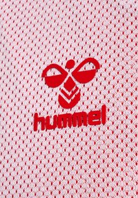 Logo rosso di Hummel su tessuto bianco, testurizzato, in rete con piccole perforazioni. Presenta un distintivo emblematico di un'ape con ali stilizzate.