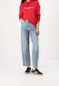 Rode hoodie met witte tekst, lichtblauwe wijde jeans met hoge taille, en zwarte sneakers met witte strepen en een gumzool.