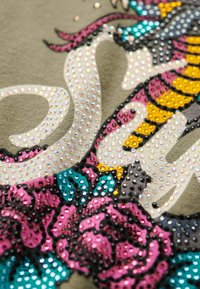 Olivengroen stof met kleurrijke rhinestone versieringen, voorzien van bloempatronen in roze, turquoise en geel, samen met een gebogen tekstontwerp.