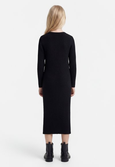 Blond persoon die een zwarte geribbelde lange mouwen midi-jurk en zwarte enkellaarsjes draagt, staand met de rug naar voren tegen een witte achtergrond.