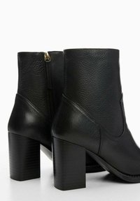 Par de botas de cuero negro por el tobillo con tacones de bloque y cremalleras laterales, acabado texturizado y liso, exhibidas sobre un fondo blanco.