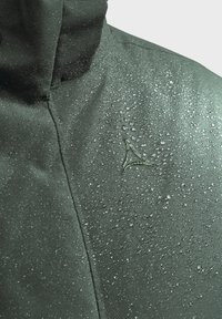 Wasserabweisender grüner Jackenstoff mit Wassertropfen, versehen mit einem dezenten schrägen Logo auf der oberen linken Seite. Glatte Textur, klares Design.