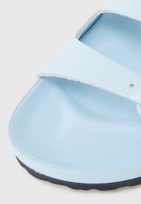 Gros plan d'une sandale bleu clair avec une large sangle et une semelle moulée, montrant les bords texturés et la semelle noire.