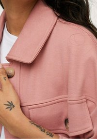 Różowa dżinsowa kurtka z dużym kołnierzem, zapięciem na guziki i przednimi kieszeniami. Tkanina ma teksturowane wykończenie z widocznymi detalami szycia.