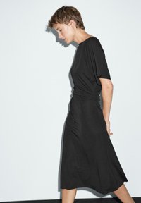 Vestito nero al ginocchio realizzato in tessuto lucido, caratterizzato da una vestibilità comoda, maniche corte e dettaglio arricciato sui fianchi per un tocco di texture in più.
