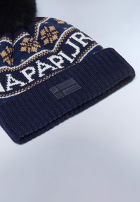 Berretto lavorato a maglia in navy e crema con motivi geometrici, caratterizzato da un pompon in pelliccia sintetica e una toppa nera rettangolare con un design a bandiera.