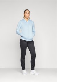 Svetlomodrá mikina na zips s kapucňou, v kombinácii s čiernymi jogger nohavicami. Biely tenisky dopĺňajú outfit, ktorý vystihuje neformálny športový štýl.