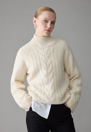 Cremfarbener Kabelstrickpullover mit hohem geripptem Kragen und weit geschnittenen Ärmeln, kombiniert mit einem weißen Hemd und schwarzen Hosen. Sanfte Textur, gemütliches Design.
