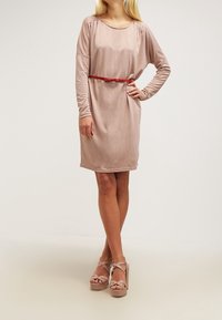 Langarmige jurk tot de knie van zacht beige stof, met een aansluitende taille en een rode riem. Gemaakt in combinatie met beige plateausandalen.