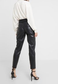 Zwarte neplederen broek met een hoge taille, voorzien van een gladde textuur, smal toelopende pasvorm en achterzakken, gecombineerd met een lichtblouse en hakken.