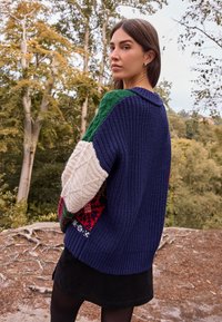 Džemper od pletenine u kombinaciji mornarsko plave, zelene, bijele i uzorkom naglašenih detalja; ima rebrastu teksturu i ležeran kroj s okruglim izrezom.