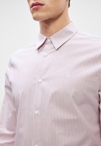 Lichtroze shirt met verticale strepen, met knoopsluiting en knoopjes aan de voorkant. Beschikt over een klein geborduurd logo aan de linkerkant.