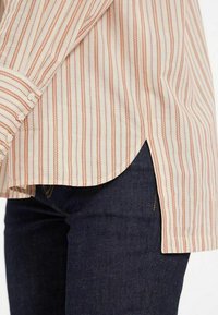Chemise rayée en crème et rouille, dotée de poignets avec boutons, d'un ourlet incurvé et d'un tissu léger. Associée à un denim bleu foncé.