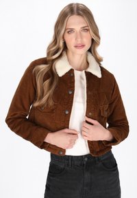 Giacca di corduroy marrone con un colletto in pelliccia sintetica color crema, tasca frontale e bottoni metallici; indossata sopra una maglietta bianca e jeans neri.