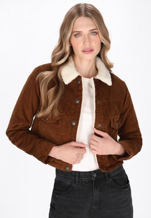 Giacca di corduroy marrone con un colletto in pelliccia sintetica color crema, tasca frontale e bottoni metallici; indossata sopra una maglietta bianca e jeans neri.