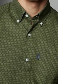 Green button-up shirt with a repeating palm leaf pattern, made of fabric with a smooth texture; features light buttons and a small logo tag.