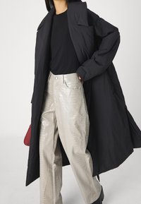 Abrigo negro ligero con cuello alto, combinado con pantalones plateados brillantes con estampado de cocodrilo. Bolso rojo visible en la cadera.